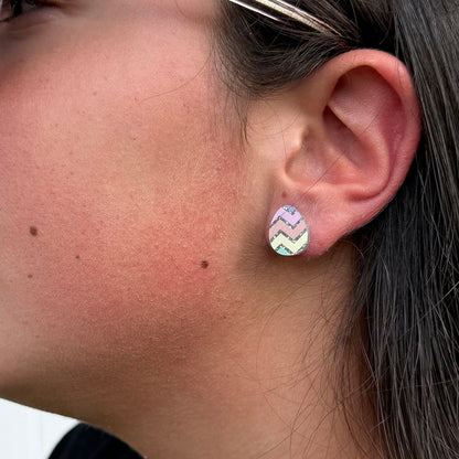 A close-up of a person's ear wearing a small Easter egg shaped stud earring with a pastel chevron pattern in pink, yellow, green, and blue.