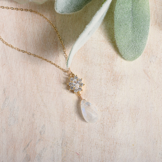 Delicate gold necklace with a cubic zirconia pendant and moonstone drop on a textured beige surface.