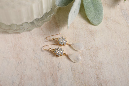 Gold earrings with CZ stones and moonstone on a beige surface with green leaves