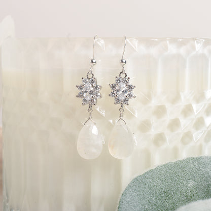 Silver earrings with clear stones and teardrop moonstone on a textured white surface.