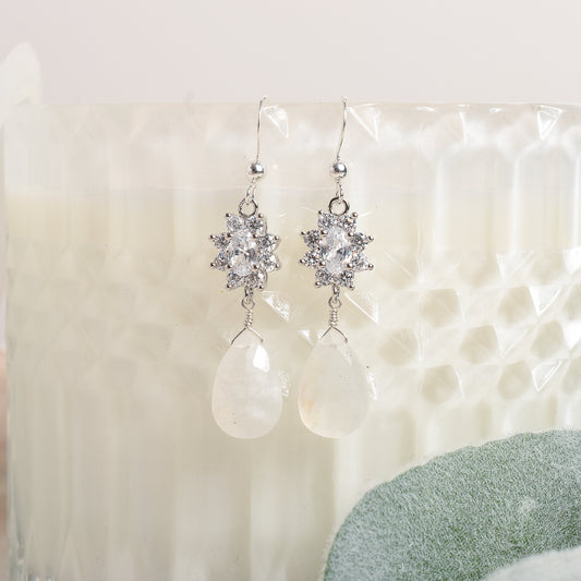 Silver earrings with clear stones and teardrop moonstone on a textured white surface.
