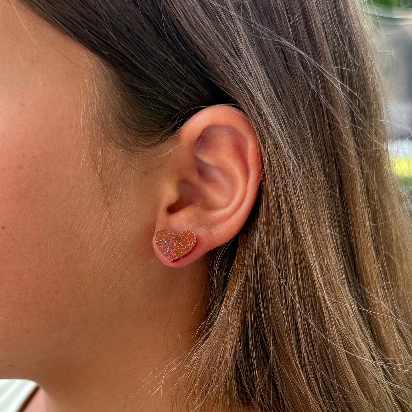 A close-up side profile of a woman's ear wearing a small glittery heart-shaped stud earring in a deep red.