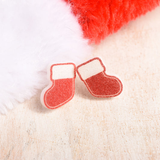 A pair of Christmas stocking-shaped stud earrings made of glittery red acrylic with white cuffs, displayed on a light wood surface next to a red and white Santa hat.