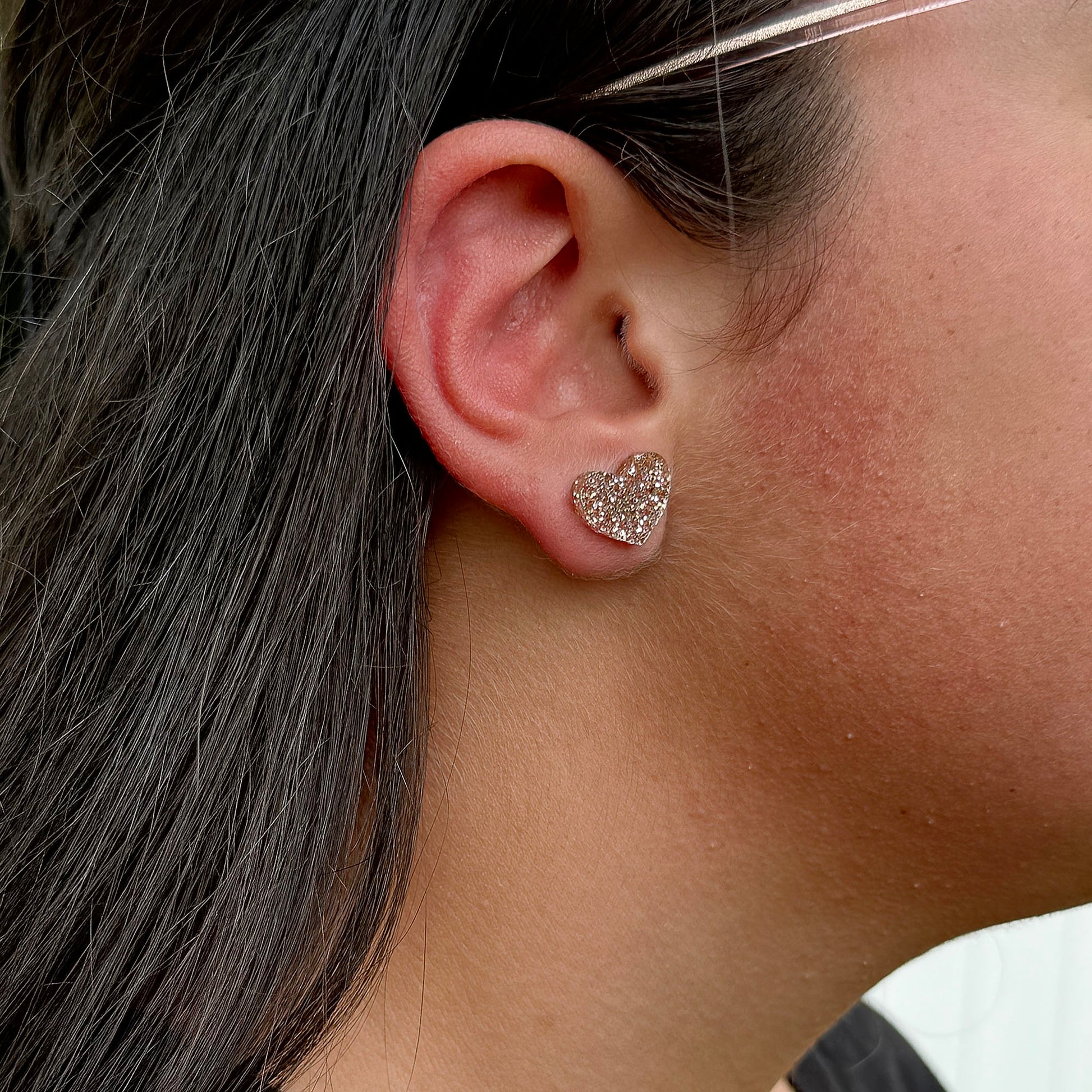 A close-up of a person's ear wearing a small heart-shaped stud earring covered in a glittery rose gold color.