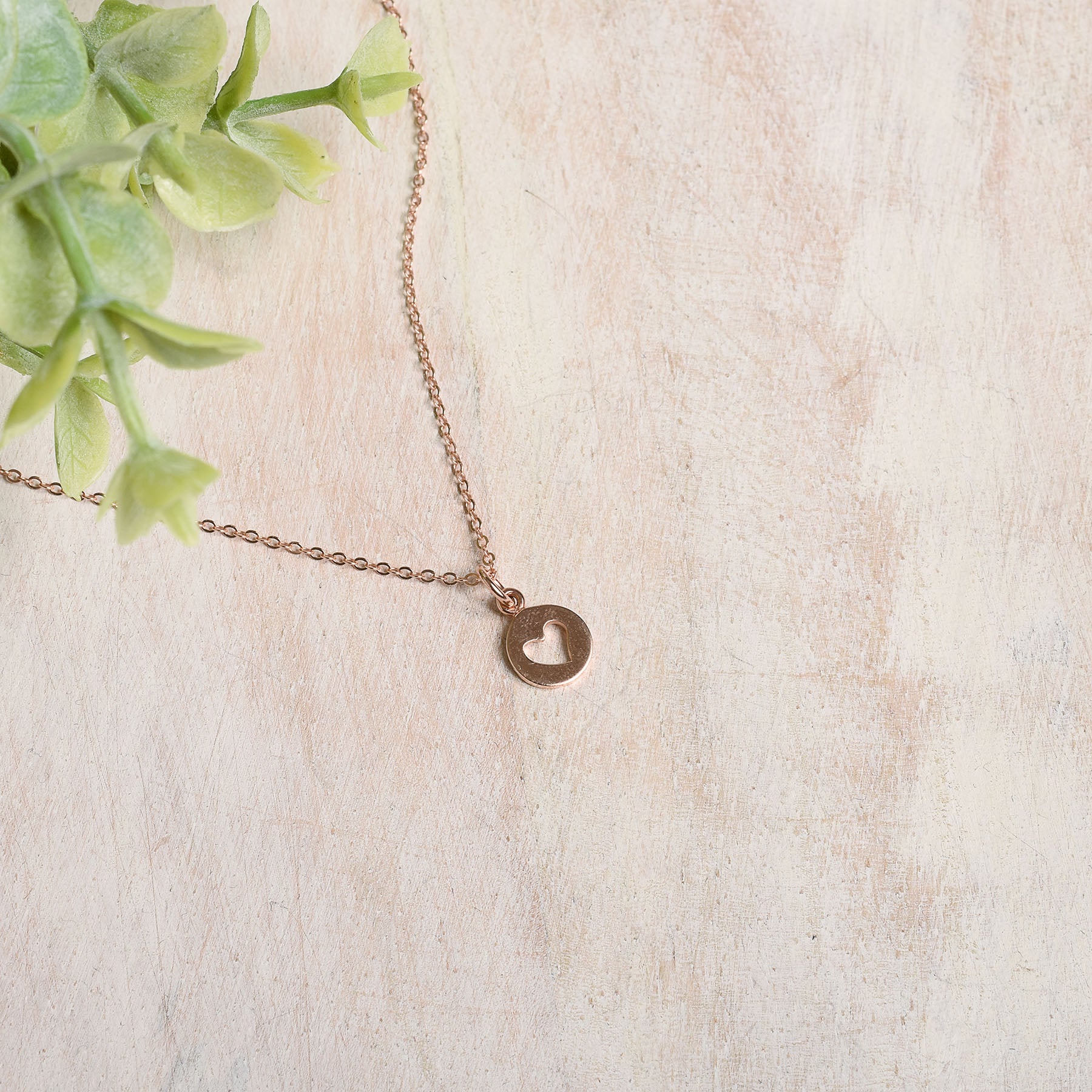 Rose gold necklace with a heart pendant on a light beige surface with green leaves.