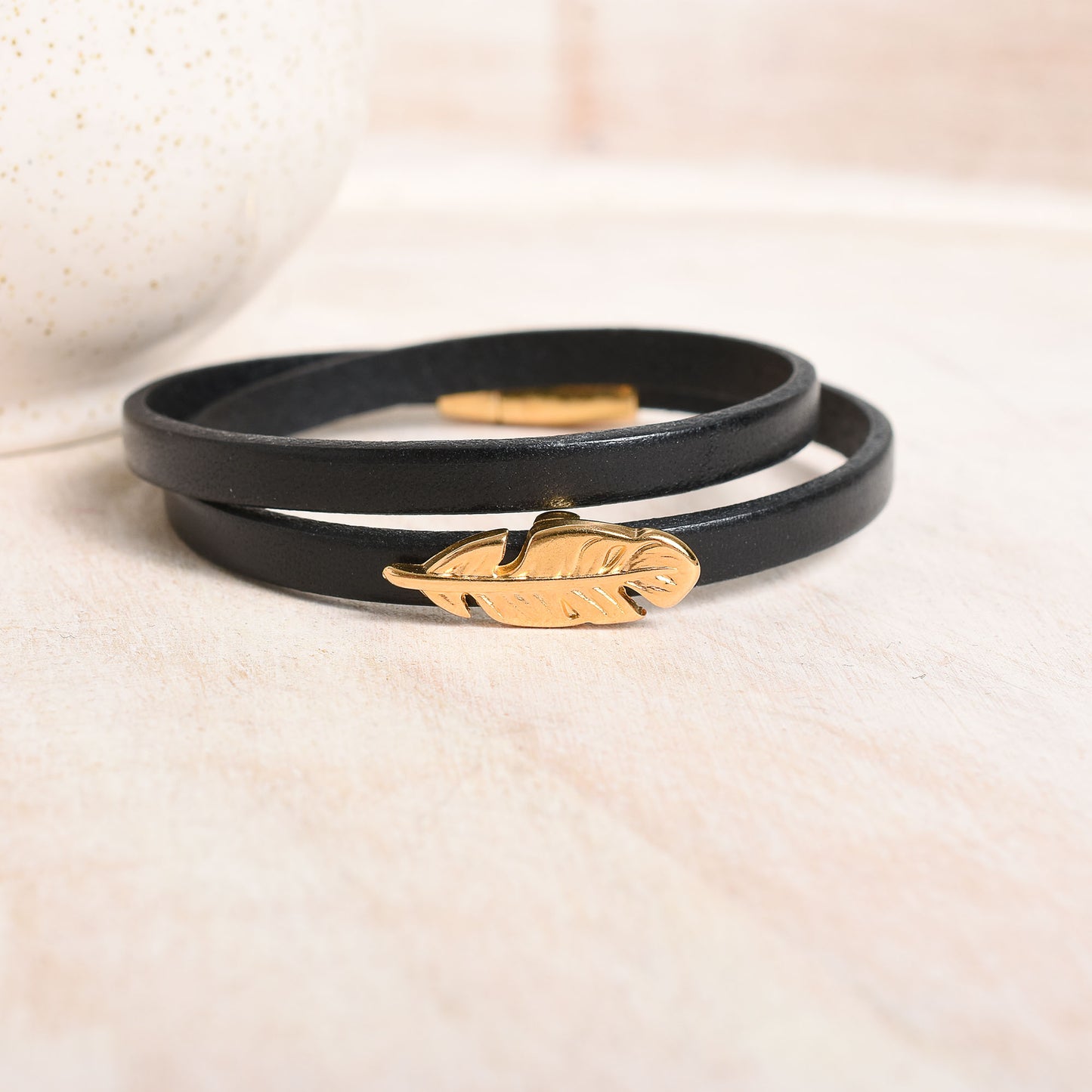 Black leather bracelet with gold feather accent on a light beige background