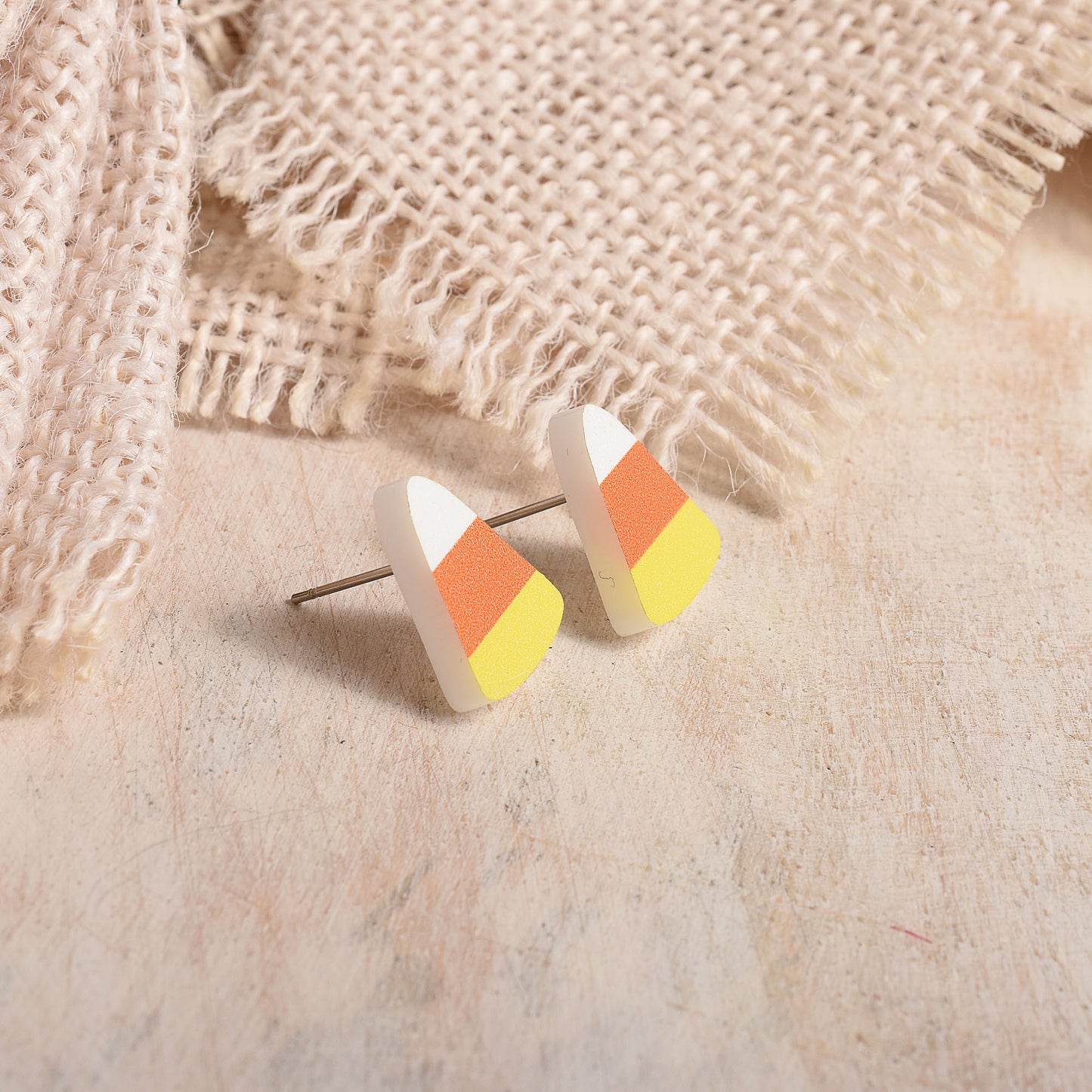 Candy corn stud earrings with white, orange, and yellow sections, displayed on a textured beige surface with burlap fabric in the background.
