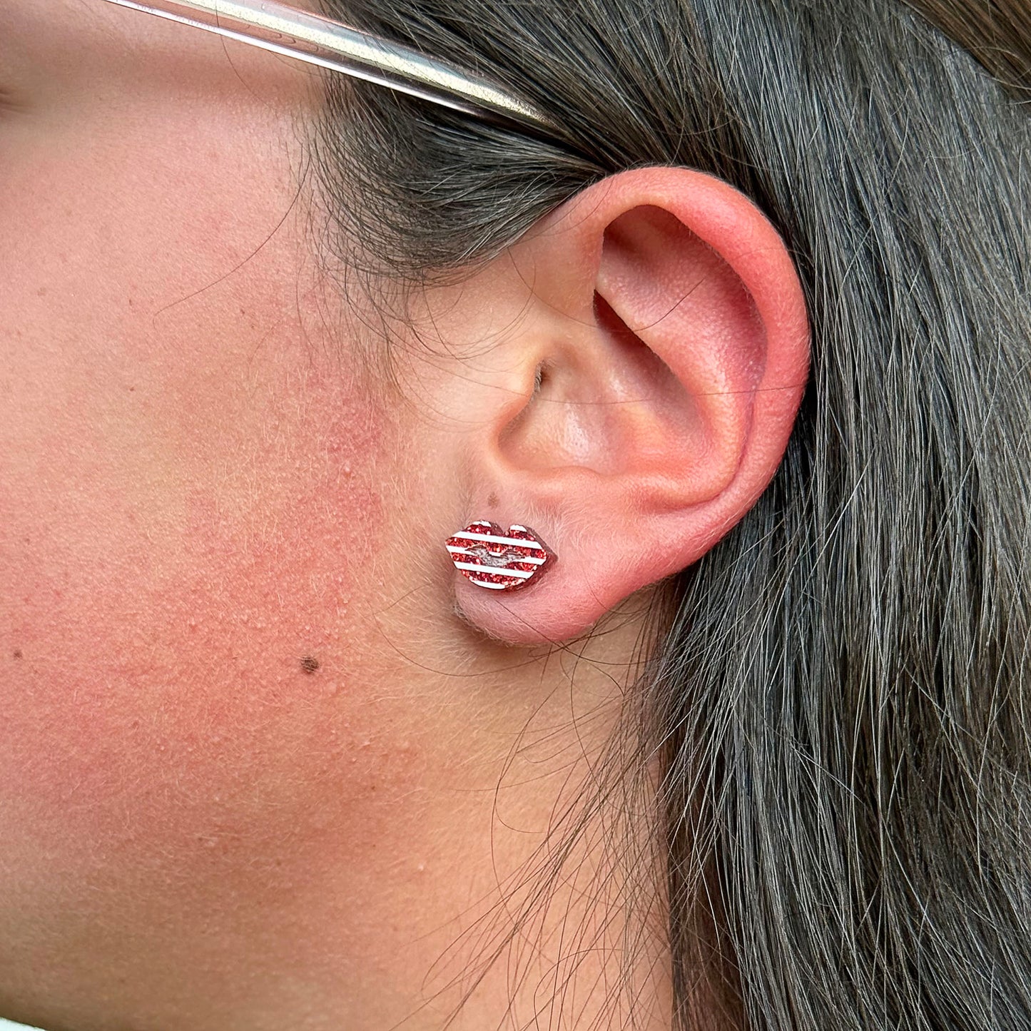 A close-up of a person's ear wearing a small lip-shaped stud earring with red and white horizontal stripes.
