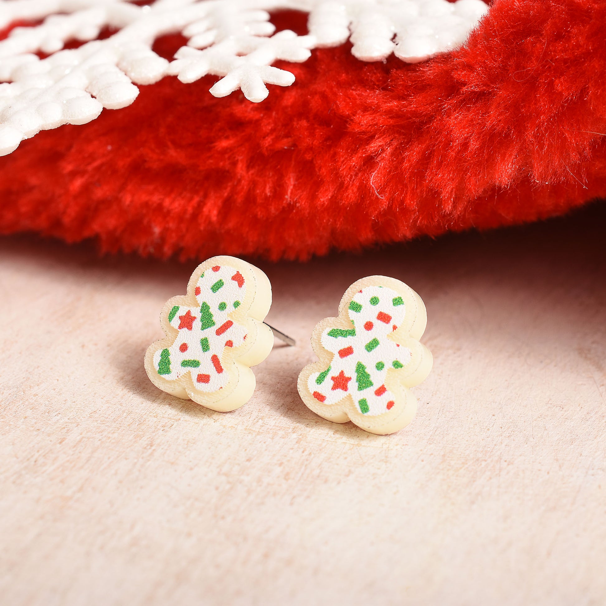 A pair of small gingerbread man stud earrings with white icing-like bodies and colorful holiday sprinkles, placed on a beige surface with a red fuzzy decorative prop in the background.