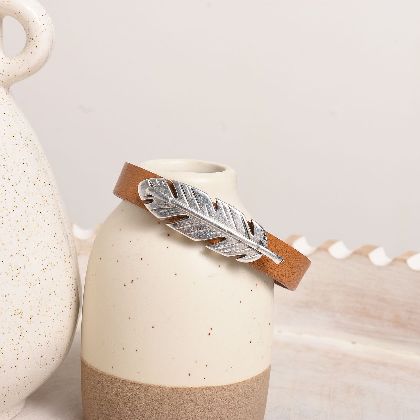 A tan leather cuff bracelet with a textured silver feather charm.