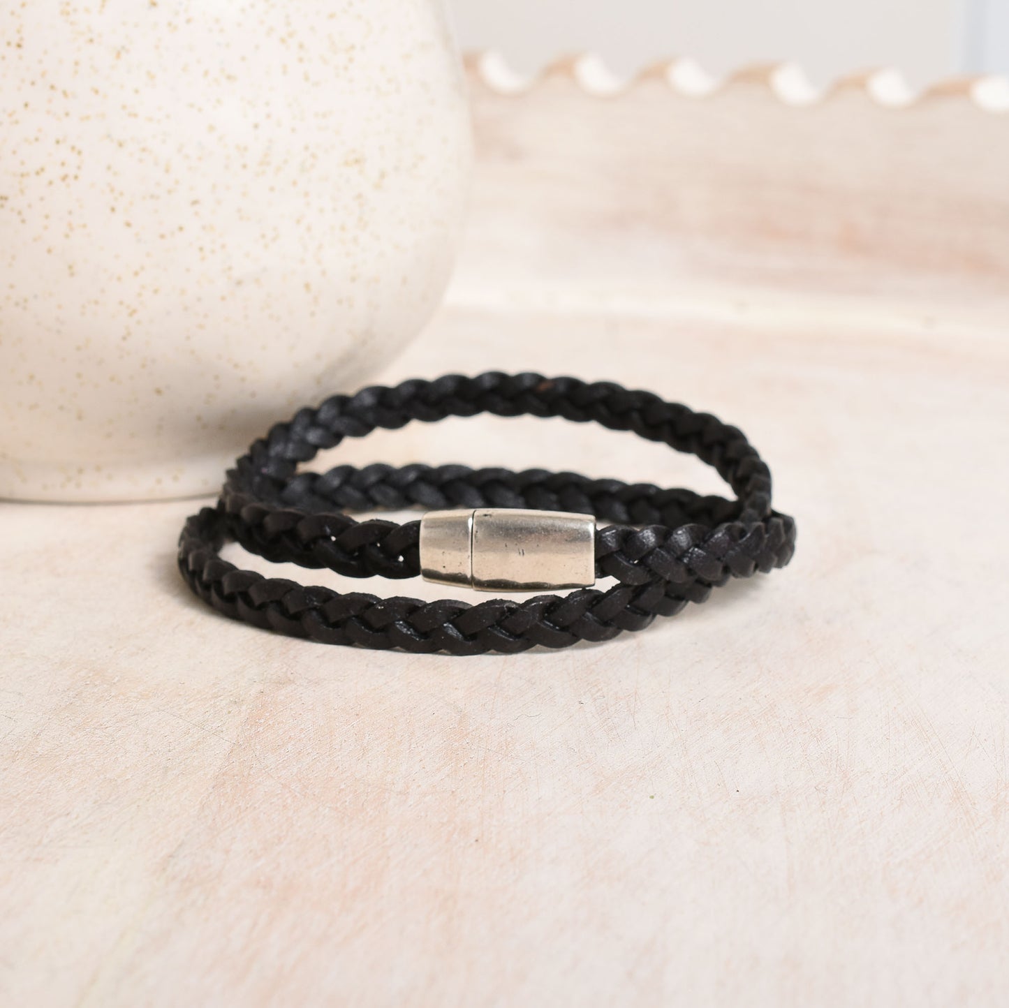 A black braided leather bracelet with a polished silver metal clasp, resting on a light beige textured surface.