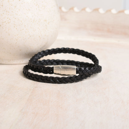A black braided leather bracelet with a polished silver metal clasp, resting on a light beige textured surface.