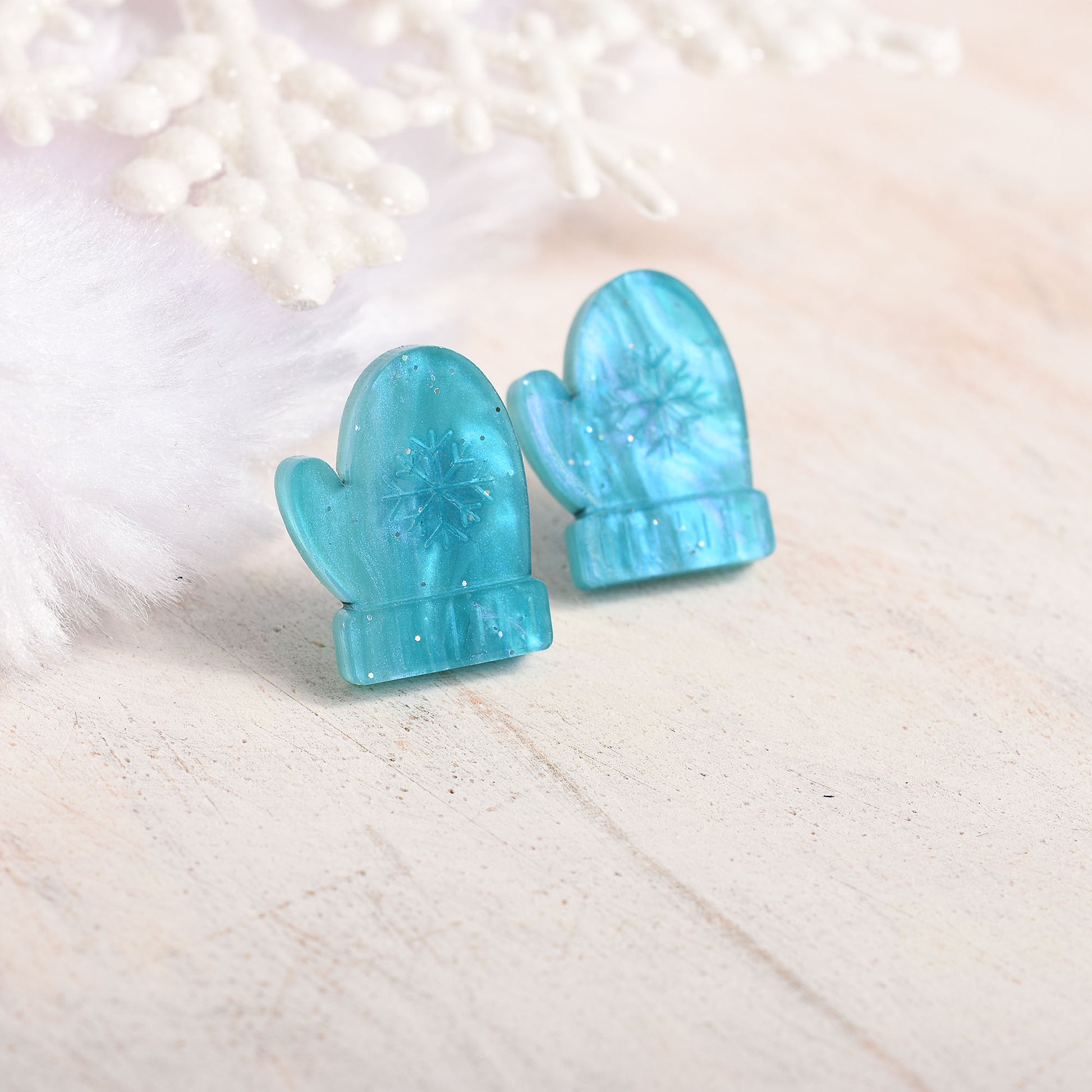 A pair of aqua blue mitten-shaped stud earrings with glitter and an embossed snowflake design, displayed on a light beige textured surface with white decorative props.