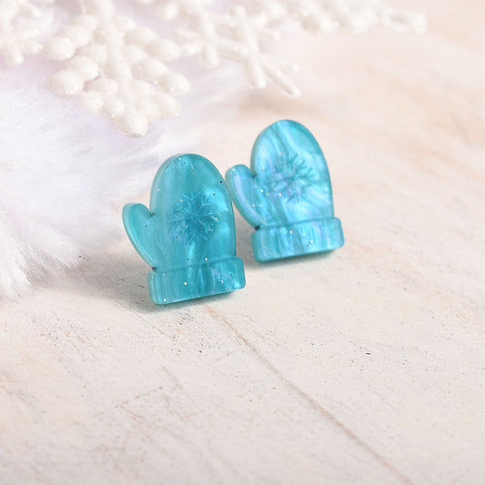 A pair of aqua blue mitten-shaped stud earrings with glitter and an embossed snowflake design, displayed on a light beige textured surface with white decorative props.