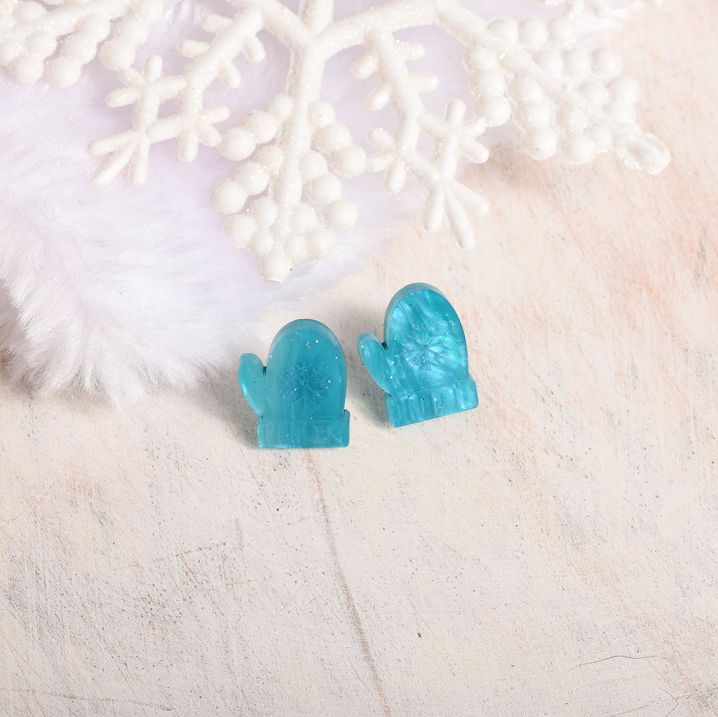 A pair of small mitten-shaped stud earrings in translucent aqua blue, displayed on a light textured surface with white decorative snowflake and faux fur props.