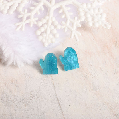 A pair of small mitten-shaped stud earrings in translucent aqua blue, displayed on a light textured surface with white decorative snowflake and faux fur props.